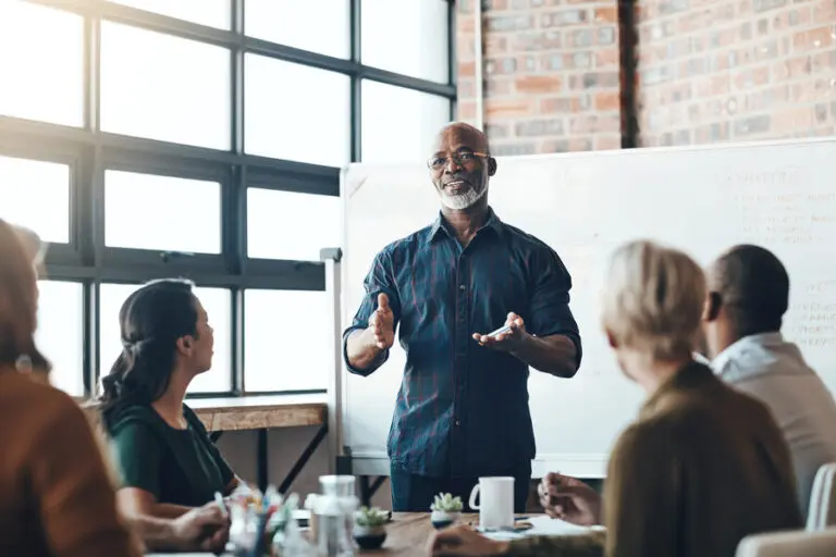 A diverse group of professionals is engaged in a meeting in a modern office setting. A senior man is standing and speaking in front of a whiteboard, gesturing with his hands as he addresses the seated colleagues who are attentively listening. The background features large windows allowing natural light to fill the room, and the office has a brick wall, giving it an industrial yet contemporary feel. The whiteboard behind the speaker has notes and diagrams relevant to the discussion.