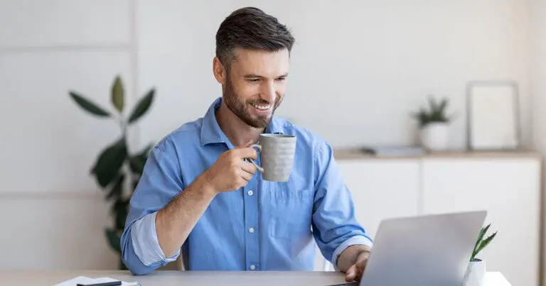 Photo of a white man with brown hair, a mustache, and beard wearing a blue button up collared shirt smiling and holing a cup of coffee while looking at a laptop in front of him