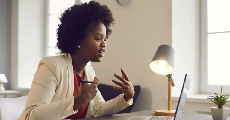 Photo of a black woman in an off-white blazer sitting at a table and speaking through a computer for an interview