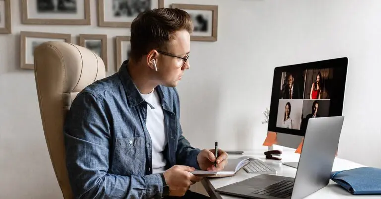 A man is seated at a desk in a home office setting, participating in a virtual meeting on his computer. He wears glasses and white earbuds, and is taking notes in a notebook. On the computer screen, there are four people visible in the video call, all dressed in professional attire. The desk is organized with a laptop, a few office supplies, and a closed notebook. The background includes framed pictures on the wall.